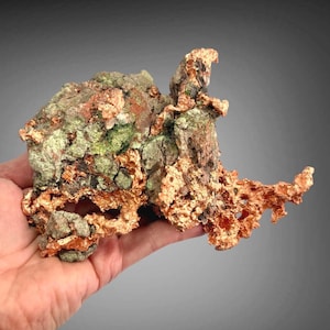 May include: A raw mineral specimen featuring a combination of metallic copper deposits and textured, light green rock formations. The piece displays a jagged, irregular shape with porous sections and a mix of shiny copper and matte green surfaces.
