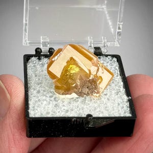 May include: A translucent, golden-yellow wulfenite crystal specimen with sharp, tabular edges and a small cluster of brown matrix attached. The mineral is displayed inside a small, square, black plastic box with a clear hinged lid.