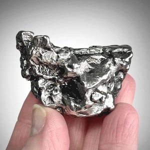 May include: A metallic, silver-coloured meteorite fragment with a highly irregular, textured surface featuring deep crevices, sharp ridges, and a reflective, polished appearance. The object has a jagged, multi-faceted shape with dark shadows.