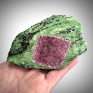 Peut inclure: Un spécimen minéral brut présentant un grand cristal magenta de forme carrée incrusté dans une matrice vert clair texturée. La pierre verte présente des inclusions minérales noires proéminentes et des stries sur toute sa surface, créant un contraste élevé.