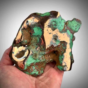 May include: A raw copper specimen featuring a mix of polished metallic surfaces and natural oxidised areas. The piece displays a vibrant contrast between shiny, smooth copper sections and textured, crusty patches of bright green malachite.