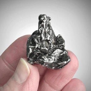 May include: A small, metallic, silver-toned object with a highly irregular, craggy, and textured surface. The piece features deep crevices, raised ridges, and a reflective, polished finish across its complex, multi-faceted, and organic shape.