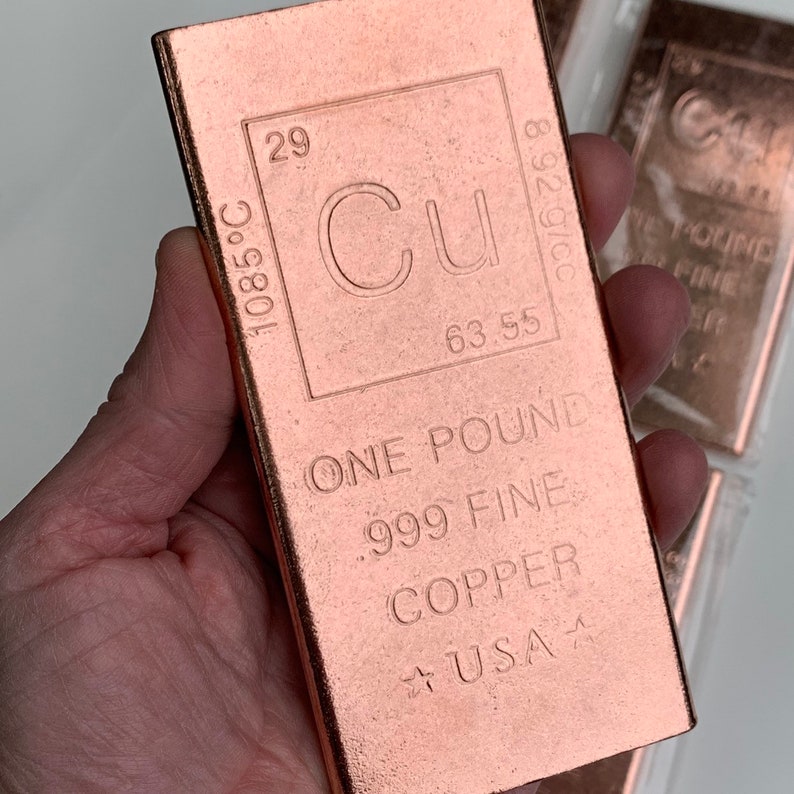May include: A copper bar with the element symbol Cu, atomic number 29, and a weight of one pound. The bar is stamped with the text "ONE POUND .999 FINE COPPER USA". The bar is held in a hand.