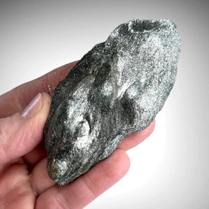 Könnte beinhalten: Ein rohes, unregelmäßiges Stück Hämatit-Mineral mit einem metallischen, silbergrauen Glanz. Die Oberfläche weist eine faserige, kristalline Textur mit reflektierenden Highlights und dunklen, anthrazitfarbenen Bereichen auf seiner unebenen Form auf.