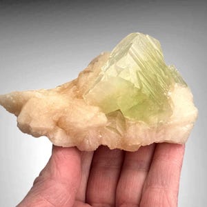 Op de afbeelding: Een lichtgroen en beige mineraal specimen. Het belangrijkste kenmerk is een grote, kristallijne formatie van lichtgroene kleur, geplaatst op een basis van beige gekleurd gesteente. Het mineraal wordt in een hand gehouden.