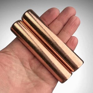 May include: Two cylindrical bars with a polished, metallic copper finish. Each bar features a smooth, rounded surface with a reflective, warm reddish-brown tone. The objects are solid, elongated, and have a uniform, straight shape throughout their length.