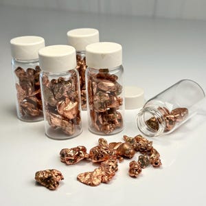 Op de afbeelding: Verschillende kleine glazen flesjes gevuld met koperklompjes, sommige met witte deksels. De klompjes hebben een glanzende, roodbruine kleur. Enkele klompjes liggen buiten de flesjes. De afbeelding is goed belicht en toont de textuur en kleur van het koper.