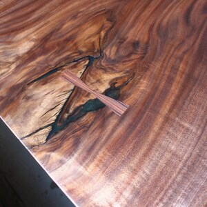 May include: A close-up of a wooden table top with a natural wood grain pattern. The table top has a dark brown colour with a lighter brown wood grain pattern. There is a small, dark brown wooden inlay in the shape of a cross in the centre of the table top.