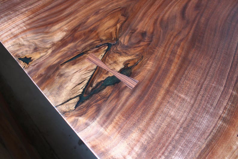May include: A close-up of a wooden table top with a natural wood grain pattern. The table top has a dark brown colour with a lighter brown wood grain pattern. There is a small, dark brown wooden inlay in the shape of a cross in the centre of the table top.