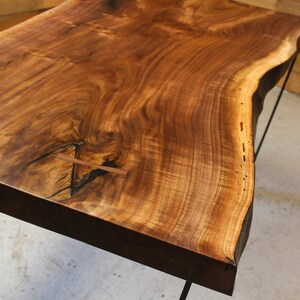 May include: A large, live edge wooden table top with a natural wood finish. The table top has a unique, irregular shape and is supported by a black metal frame.