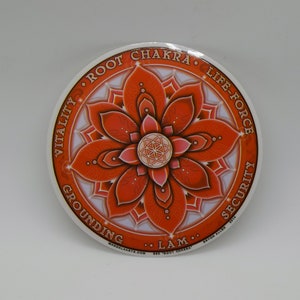 Puede incluir: Una pegatina redonda con un diseño de mandala naranja y blanco. El mandala presenta una flor con ocho pétalos y un diseño geométrico más pequeño en el centro. El texto alrededor del mandala dice "Root Chakra", "Life-Force", "Security", "Grounding", "Vitality" y "Lam".