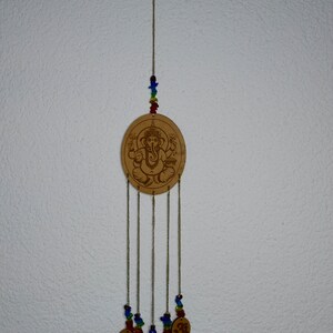 May include: A wooden wind chime with a circular design featuring an image of the Hindu deity Ganesha. The chime has five strands of beads and five small circular charms with the Om symbol.