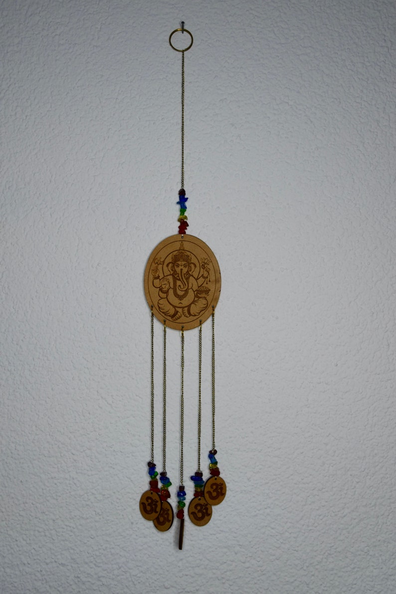 May include: A wooden wind chime with a circular design featuring an image of the Hindu deity Ganesha. The chime has five strands of beads and five small circular charms with the Om symbol.