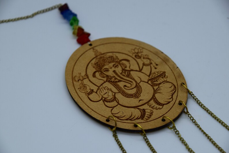 May include: A wooden wall hanging with a carved image of the Hindu deity Ganesha. The hanging has a chain with colorful beads and four chains hanging from the bottom.