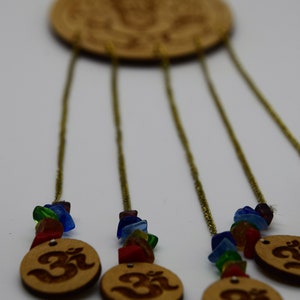 May include: A wooden wind chime with five strands of beads and a circular wooden top with an Om symbol. The beads are made of colorful glass and are strung on gold chains. The Om symbol is carved into the wood and is visible on the top of the chime.