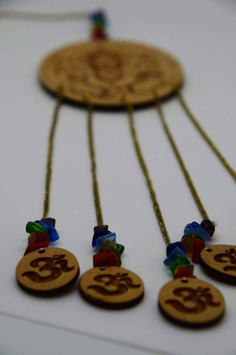 May include: A wooden wind chime with five strands of beads and a circular wooden top with an Om symbol. The beads are made of colorful glass and are strung on gold chains. The Om symbol is carved into the wood and is visible on the top of the chime.