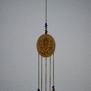 May include: A wooden wind chime with a circular design featuring an image of the Hindu deity Ganesha. The chime has five strands of beads and five charms with the Om symbol.