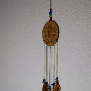May include: A wooden wind chime with a brown oval charm featuring a carved image of Ganesha. The charm is suspended from a string of colorful beads and hangs from a chain with five smaller charms featuring the Om symbol.