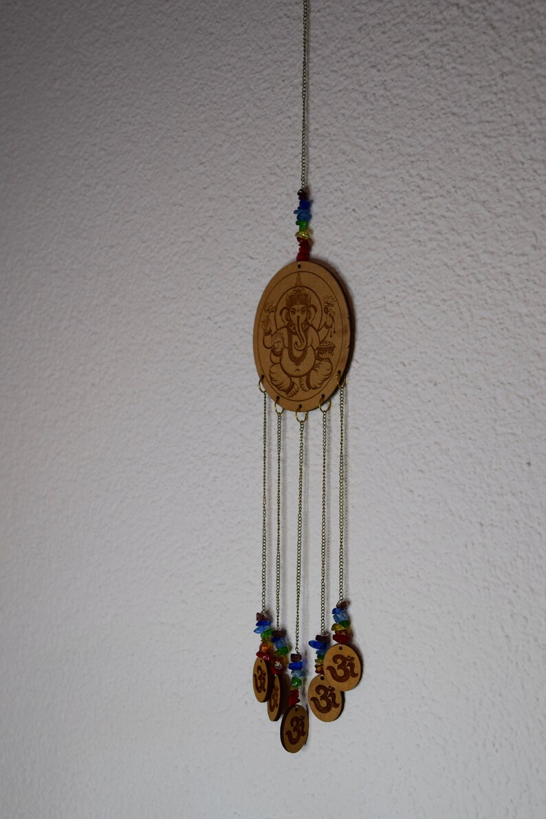 May include: A wooden wind chime with a brown oval charm featuring a carved image of Ganesha. The charm is suspended from a string of colorful beads and hangs from a chain with five smaller charms featuring the Om symbol.