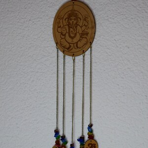 May include: A wooden wind chime with a circular cutout featuring an image of the Hindu deity Ganesha. The chime has five strands of beads and charms, including Om symbols, hanging from the bottom.