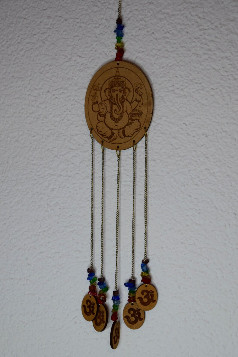 May include: A wooden wind chime with a circular cutout featuring an image of the Hindu deity Ganesha. The chime has five strands of beads and charms, including Om symbols, hanging from the bottom.