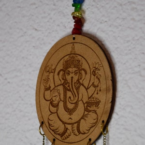 May include: A wooden oval-shaped wind chime with a carved image of the Hindu deity Ganesha. The chime has five chains with rings at the bottom. A string of colorful beads hangs from the top of the chime.