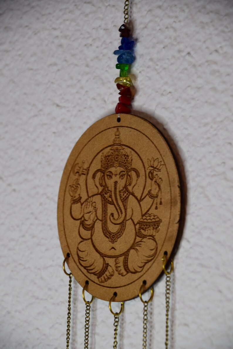 May include: A wooden oval-shaped wind chime with a carved image of the Hindu deity Ganesha. The chime has five chains with rings at the bottom. A string of colorful beads hangs from the top of the chime.
