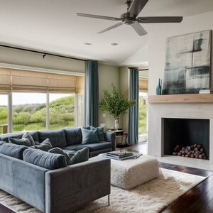 May include: A large, sectional, blue sofa with white throw pillows sits in front of a large window with a view of green hills. A white ottoman sits in front of the sofa. A fireplace with a white mantel and a black firebox is on the right side of the image. The fireplace is decorated with a black and white abstract painting.