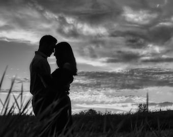 May include: A black and white silhouette of a couple embracing under a cloudy sky. The couple is standing in a field of tall grass.