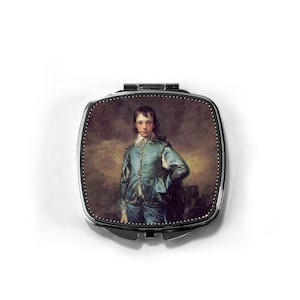 May include: A square, silver-toned compact mirror featuring a portrait of a young person in a blue suit. The artwork is a classic painting with a dark background and a detailed depiction of the subject's clothing and pose.