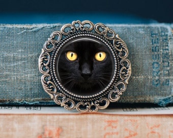 May include: A black cat with bright yellow eyes is featured in a silver-toned, ornate, circular frame. The frame has a decorative design with intricate details.