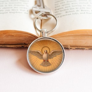 May include: A silver key chain with a round, glass charm featuring a vintage illustration of a white dove with outstretched wings, surrounded by a golden halo and rays of light.