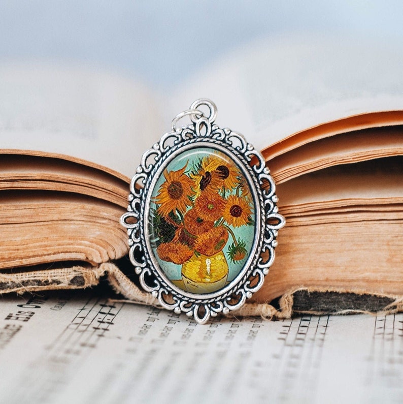 May include: A silver oval pendant with a detailed ornate frame. The pendant features a glass cameo with a painting of sunflowers in a vase. The painting is in a vintage style.