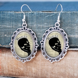 Op de afbeelding: Zilverkleurige oorbellen met een cameo-ontwerp met een zwart silhouet van een schedel met het woord "HAMLET" erboven gedrukt. De cameo is geplaatst tegen een achtergrond van vintage boekpagina's.