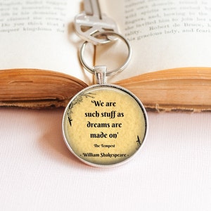 May include: A silver keychain with a round glass charm featuring a quote from Shakespeare's The Tempest: "We are such stuff as dreams are made on." The quote is printed on a vintage-style background with a floral design and birds.