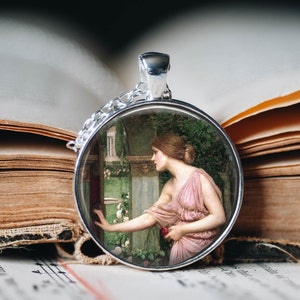 May include: A silver pendant necklace with a round glass cameo featuring a painting of a woman in a pink dress standing in a garden. The woman is looking to the left and holding a red rose.