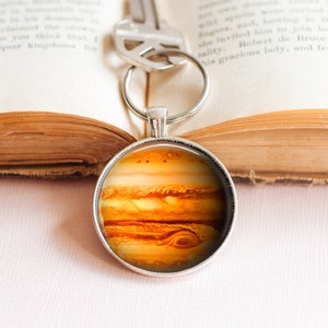 May include: A silver key chain with a round glass charm featuring a detailed image of Jupiter, the largest planet in our solar system. The planet's swirling orange and brown clouds are visible in the image.