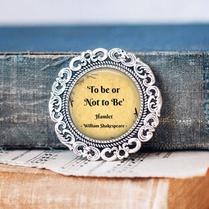 Op de afbeelding: Een rond, zilverkleurig, versierd frame met een glazen deksel omsluit een vintage-stijl papier met de quote 'To be or Not to Be' uit Hamlet van William Shakespeare.