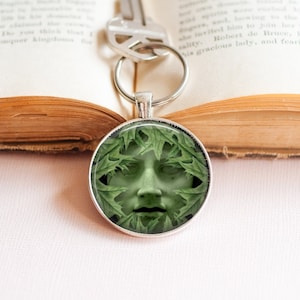 May include: A silver key chain with a round glass charm featuring a green face surrounded by leaves.