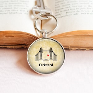 Peut inclure: Un porte-clés rond en argent avec une image de style vintage du pont suspendu de Clifton à Bristol, en Angleterre. L'image est sur un fond crème avec un cœur rouge au centre du pont. Le texte "Bristol" est imprimé sous le pont.