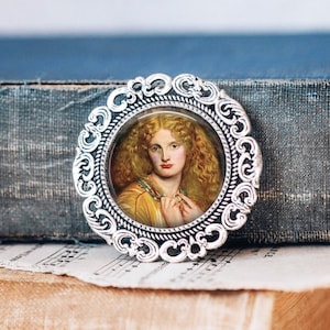 May include: A round, silver-toned brooch with an ornate, filigree-style border. The brooch features a portrait of a woman with long, curly, golden hair, wearing a yellow dress and holding a necklace. The background is blurred.
