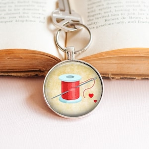 Può includere: Un portachiavi d'argento con un ciondolo rotondo che presenta un rocchetto di filo rosso, un ago e tre cuori rossi su uno sfondo in stile vintage.