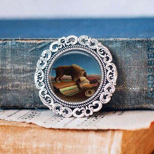 Peut inclure: Broche ronde argentée avec une bordure en filigrane ornée. Elle présente une peinture miniature d'un lion et d'une personne se reposant sur une couverture colorée, sur fond de paysage désertique.