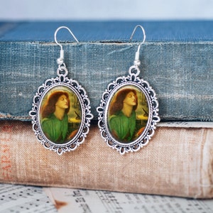 May include: Silver tone earrings with a cameo image of a woman with red hair and a green dress. The image is set in a silver oval frame.