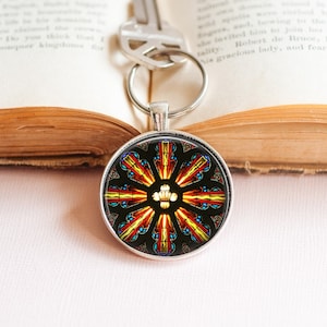 May include: A round silver keychain with a stained glass design. The design features a radial pattern of red, yellow, and blue segments against a black background. A key is attached to the keychain, resting on an open book.