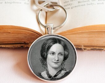 May include: A silver keychain with a round pendant featuring a black and white portrait of a woman. The keychain is resting on an open book. The portrait shows a woman with dark hair and a high-collared dress.