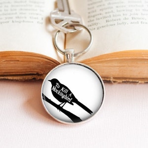 Könnte beinhalten: Ein silberner Schlüsselanhänger mit einem runden Glasanhänger, der eine schwarze Silhouette eines Spottdrossels auf einem Ast zeigt. Der Anhänger trägt den Text "To Kill A Mockingbird" in weiß.