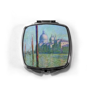 May include: A square, silver-framed compact mirror featuring a painting of a Venetian canal scene. The artwork depicts a church with a large dome, set against a blue sky and green water. The compact is closed and rests on a white background.