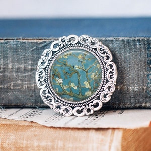 May include: A silver brooch with a round, ornate frame featuring a painting of a blooming almond tree with a blue background.