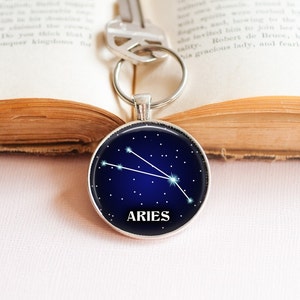 May include: A silver keychain with a round blue glass charm featuring the constellation Aries and the word "ARIES" in white lettering.